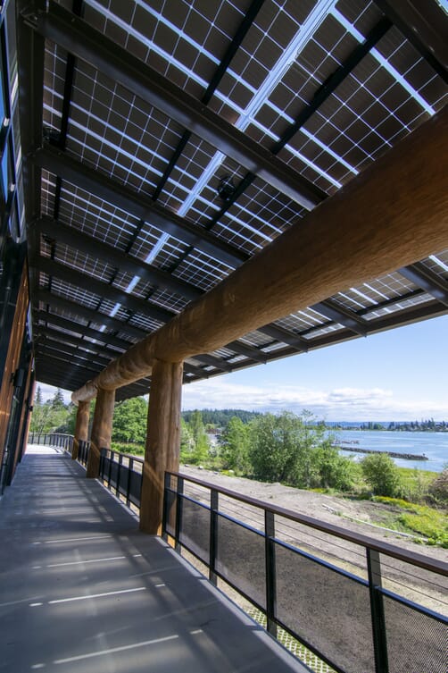 LSX Solar Panel Awning covering wraparound deck outside of Tulalip Gathering Hall with large wooden beams looking down walkway, with view of lake and trees in background