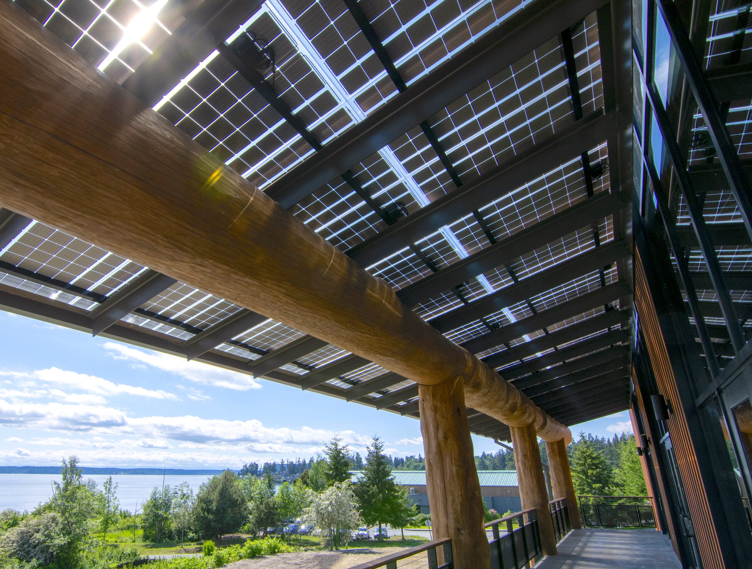LSX Solar Panel Awning covering wraparound deck outside of Tulalip Gathering Hall with large wooden beams and view of lake in background.