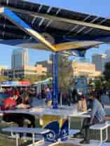 SolarZone Recharge Station with people sitting and eating on benches under solar shade and charging phone devices.