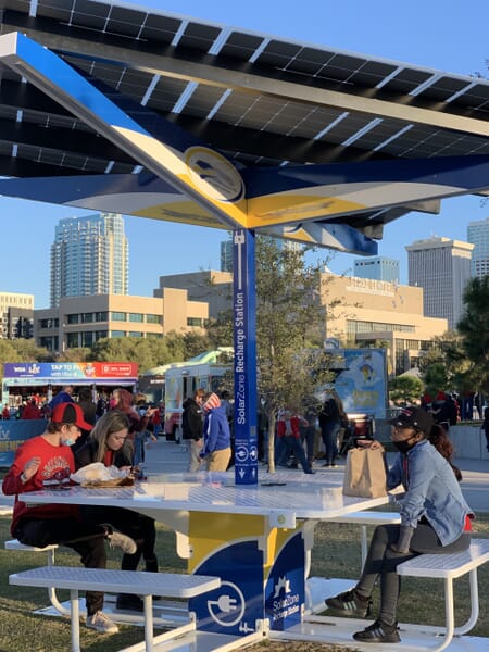 SolarZone Recharge Station with people sitting and eating on benches under solar shade and charging phone devices.
