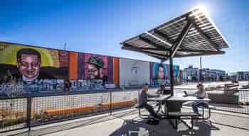 Solar Charging Table in commercial outdoor space in front of a wall mural with people sitting at benches and working on laptop computers.